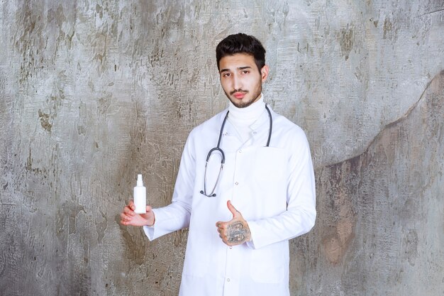 白い手の消毒剤のボトルを保持し、肯定的な手の兆候を示す聴診器を持つ男性医師。