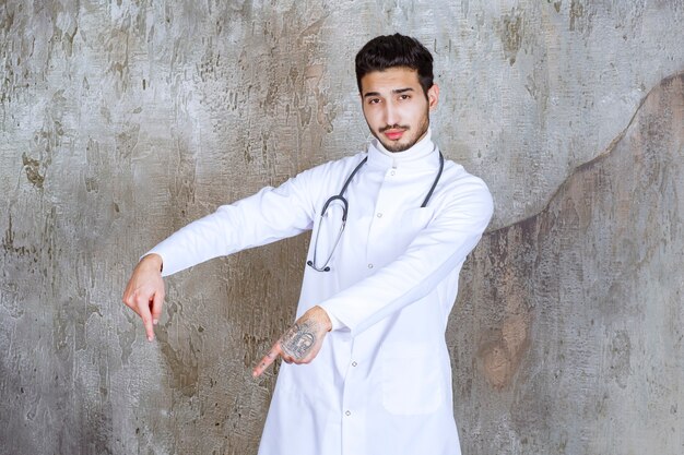 Male doctor with stethoscope standing on concrete wall and showing something below.