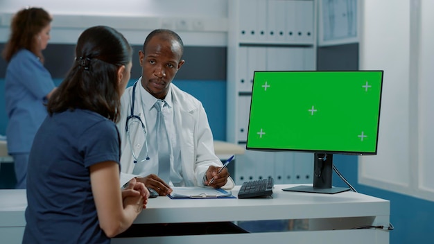 남성 의사와 여성은 의료 캐비닛에 있는 컴퓨터에서 녹색 화면을 사용합니다. 의사와 부모는 디스플레이에 격리된 복사 공간과 크로마 키 배경이 있는 빈 모형 템플릿을 보고 있습니다.