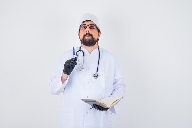 Male doctor writing while thinking in white uniform,glasses and looking concentrated , front view.