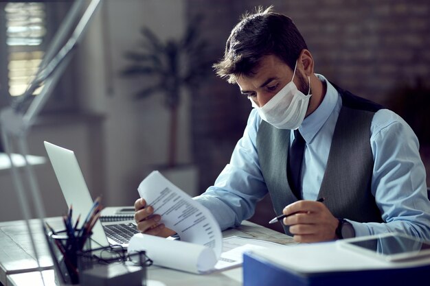 フェイスマスクを着用し、ウイルスの流行中にオフィスで働いている間、ビジネスレポートを分析する男性起業家