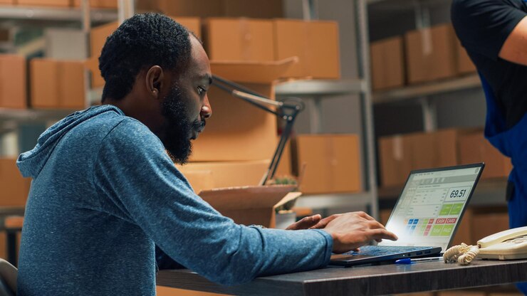 Free Photo male entrepreneur analyzing stock products and orders, doing inventory on laptop at warehouse. young man using logistics and financial planning to send merchandise for development. handheld shot.