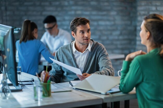 Мужчина-предприниматель обсуждает бизнес-отчеты с коллегой-женщиной во время работы за офисным столом. На заднем плане есть люди.