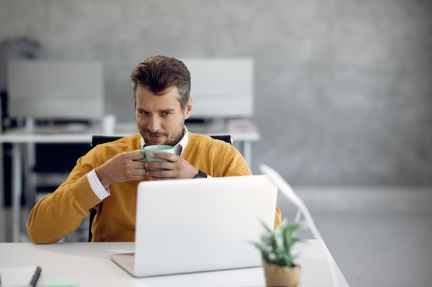 Foto gratuita imprenditore maschio che beve una tazza di caffè mentre utilizza il laptop in ufficio