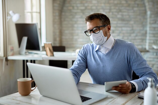 Male entrepreneur with protective face mask using touchpad while working on laptop in the office