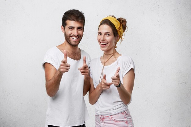 Male and female best friends with positive expression looking with pleasant smiles pointing at you with fingers having fun and good rest posing