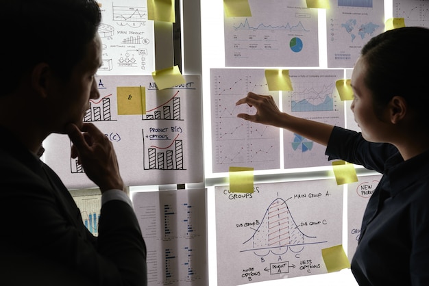 Male and female colleagues standing by window and looking at business graphs on it