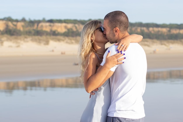 Male and female couple spending time at beach. Husband and wife in casual clothes hugging and kissing on summer day. Vacation, happiness, relationship concept