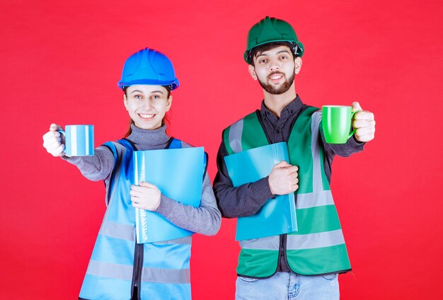 Male and female engineers with helmet holding blue and green mugs and reporting folders and making cheers.