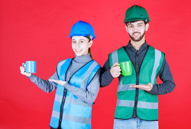 Male and female engineers with helmet holding blue and green mugs.