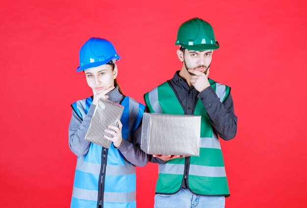 Male and female engineers with helmets holding silver gift boxes and look confused and thoughtful.