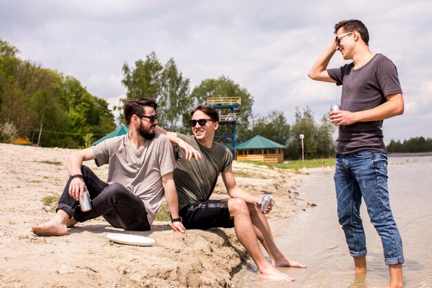 Male friends in sunglasses sitting on beach and talking 