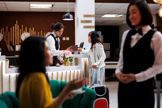 등록하기 전에 방문객의 여권을 확인하는 럭셔리 리조트의 남성 프론트 데스크 접수원. 아시아 방문객은 현대적인 호텔 예약 준비가 된 현대적인 대기 라운지에서 커피를 마시고 있습니다.