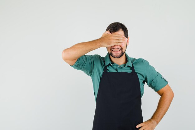 Tシャツ、エプロンで手で目を覆い、興奮している男性の庭師。正面図。