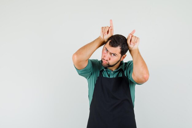 Male gardener holding fingers over head as bull horns in t-shirt, apron and looking funny , front view.