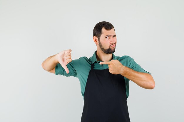 Male gardener pointing at his thumb down in t-shirt, apron and looking disappointed. front view.
