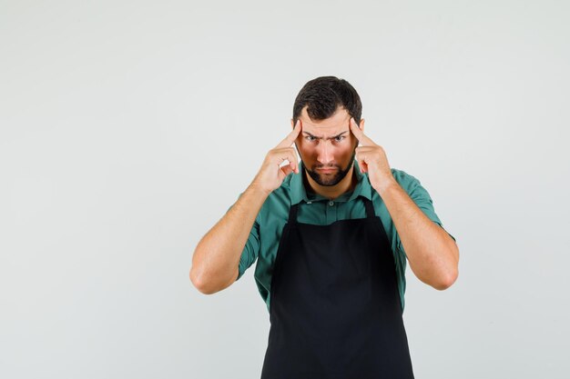 Male gardener in t-shirt, apron holding fingers on temples and looking serious , front view.