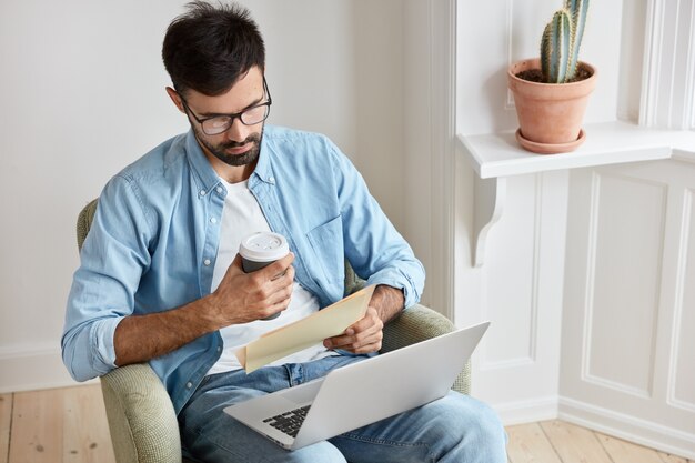 男性のグラフィックデザイナーは、ポータブルラップトップコンピューターで創造的なアイデアについてのチュートリアルビデオを見て、ビジネスニュースを読み、紙と持ち帰り用のコーヒーを持ち、自宅からフリーランスで働き、肘掛け椅子に座っています