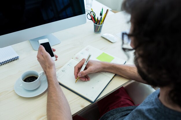 Male graphic designer writing on a diary and holding mobile phone