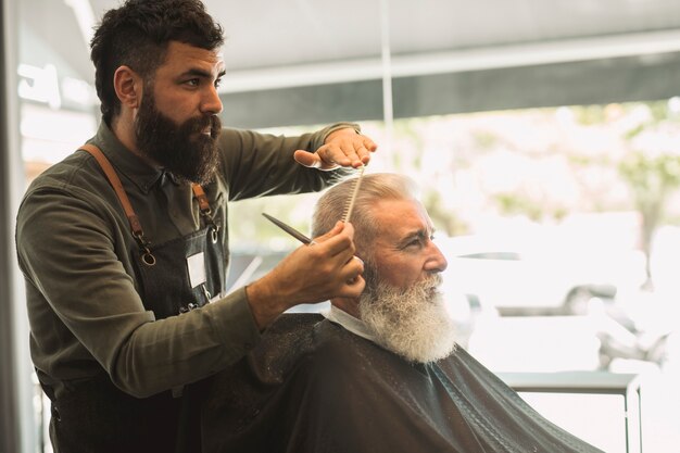 男性の美容師が理髪店で高齢者のクライアントの髪をとかす