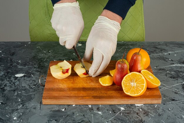 Мужская рука в перчатках, режущих свежее яблоко на мраморном столе.