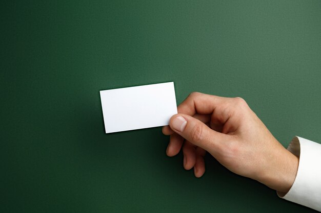テキストまたはデザインの緑の壁に空白の名刺を持っている男性の手。連絡先またはビジネスで使用するための空白のクレジットカードテンプレート。財務、オフィス。コピースペース。