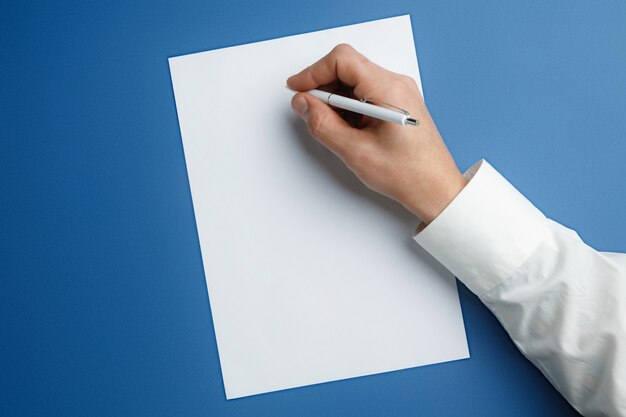 Male hand holding pen and writing on empty sheet on blue