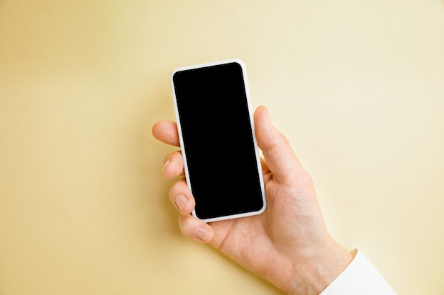 Male hand holding smartphone with empty screen on yellow wall for text or design. Blank gadget templates for contact or use in business. Finance, office, purchases.  Copyspace.