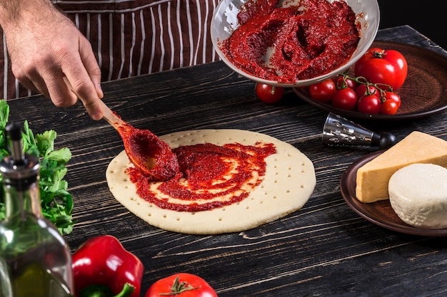 古い木製の背景にスプーンでピザベースにトマトピューレを広げる男性の手。料理のコンセプト。閉じる