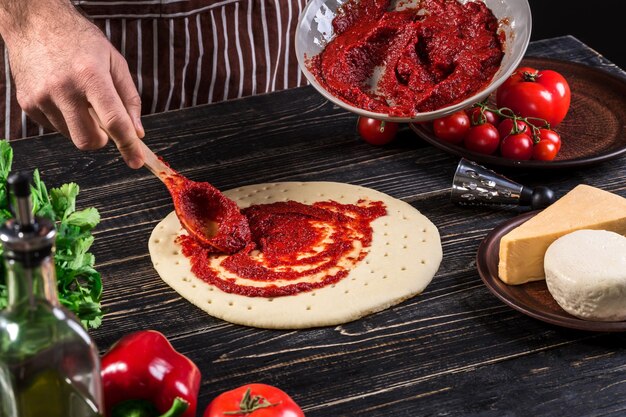 A male hand spreading tomato puree on a pizza base with spoon on an old wooden background. Cooking concept. Close-up