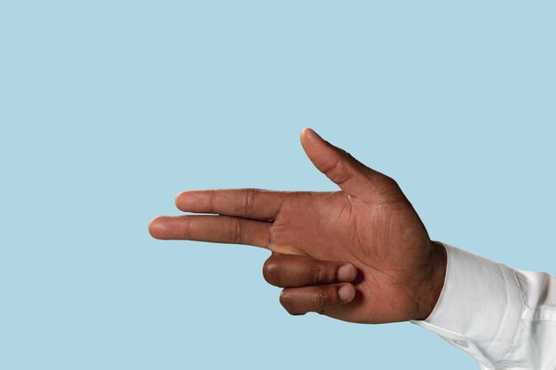 Male hand in white shirt demonstrating a gesture of gun, handgun or pistol isolated on blue wall.