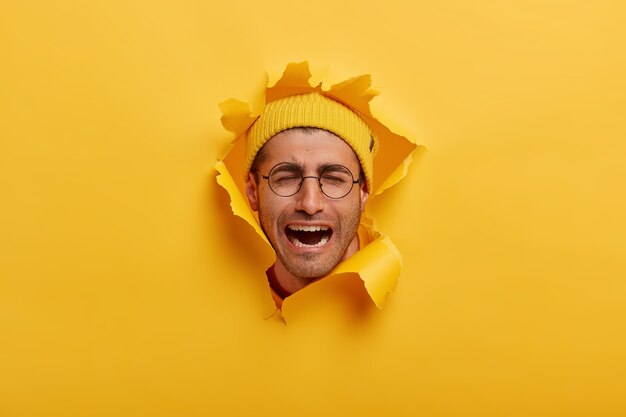 Male head in hole of paper wall. Desperate crying European man wears round spectacles and yellow hat, expresses negative emotions, keeps mouth opened