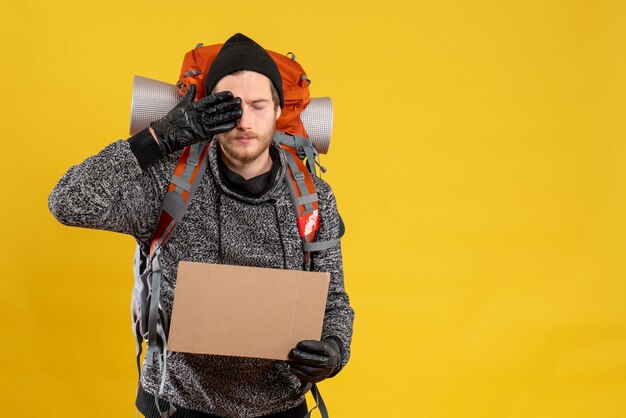 male hitchhiker with leather gloves and backpack holding blank cardboard putting hand on his eye