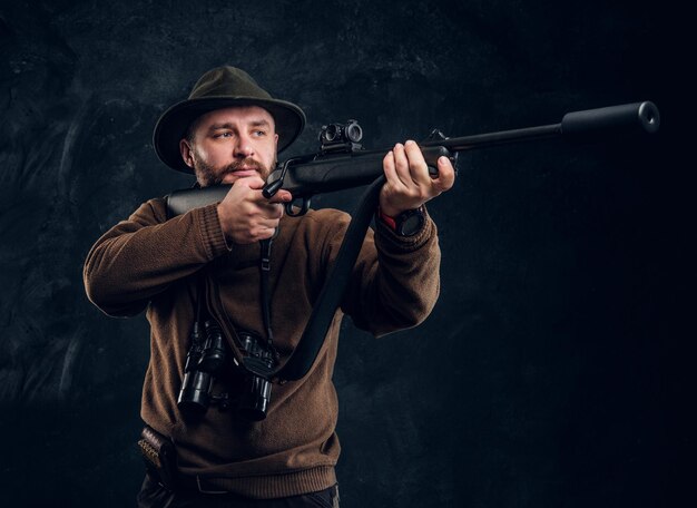 소총을 들고 그의 목표물이나 먹이를 조준하는 남성 사냥꾼. 어두운 벽 배경 스튜디오 사진
