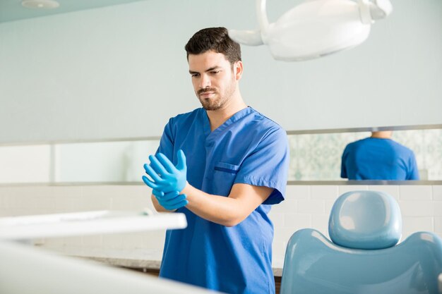 Male mid adult dentist wearing surgical gloves while standing by chair at clinic