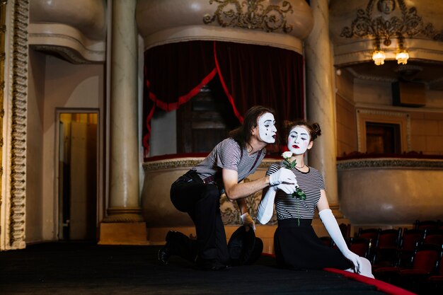 Male mime artist giving white rose to female mime sitting on stage