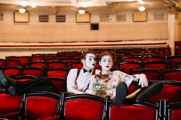 Free photo male mime holding red rose sitting with bored female mime on chair in auditorium