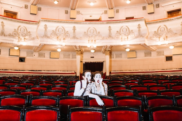 Male mime whispering in shocked female mime's ear sitting on chair in auditorium