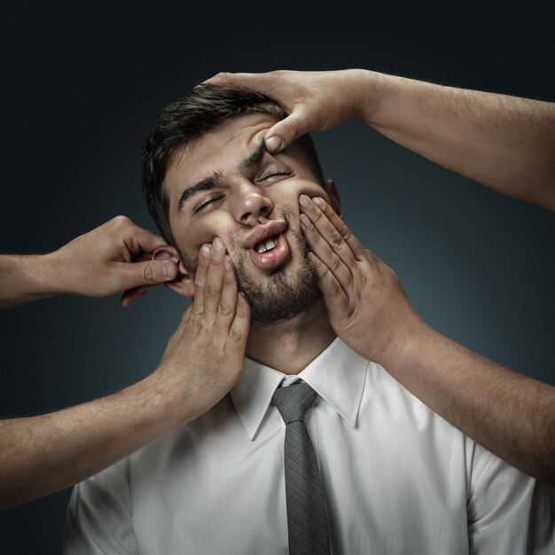 A male model surrounded by hands like his own thoughts on dark wall. A young man doubts, can't choose the right decision. Concept of mental problems, troubles in work, indecision.