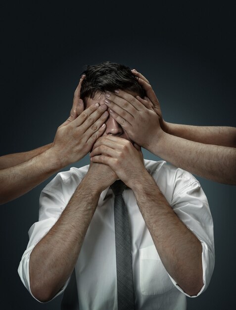 A male model surrounded by hands like his own thoughts on dark wall. A young man doubts, can't choose the right decision. Concept of mental problems, troubles in work, indecision.