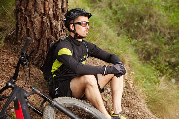 彼の折衷的な自転車で木の下の地面に座って、サイクリング旅行で休んでいる男性のマウンテンバイク