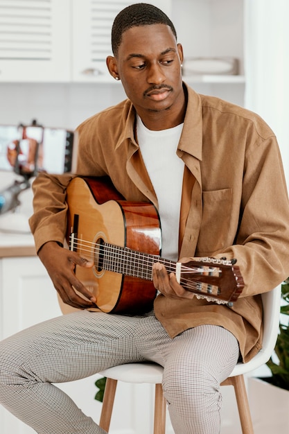 Free photo male musician at home on chair playing guitar and recording with smartphone