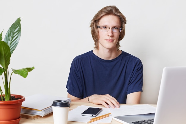 Male office worker with trendy hairstyle, wears spectacles and t shirt, sits at table, works with documents