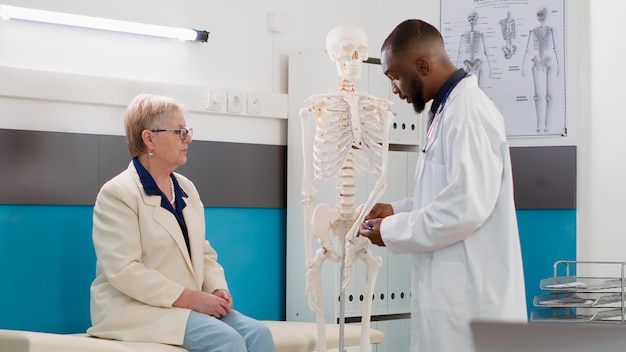 男性の整骨医は、年配の女性に人間の骨格の骨を調べ、解剖学的脊髄を分析して患者に整形外科的診断を下します。クリニックでのオステオパシー相談予約。