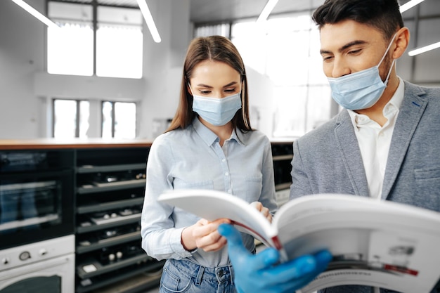 Male shop assistant in mask showing female client household appliances catalog