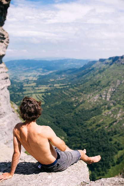 바위 절벽 위에 앉아 산과 녹지의 아름다운 전망을 즐기는 남성