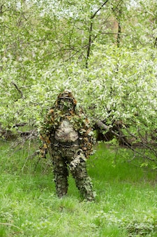 桜の近くの森で緑のカモフラージュ迷彩服でポーズをとっている男性の狙撃兵。陸軍、軍隊、エアガン、趣味の概念