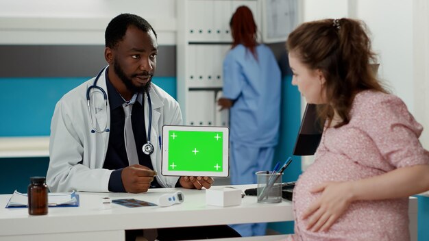 キャビネットのディスプレイに水平グリーンスクリーンとタブレットを保持している男性のスペシャリスト。空白のモックアップテンプレートで孤立したクロマキーの背景を見ている医師と妊娠中の女性。三脚ショット。