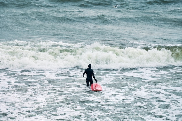 Мужчина-серфер в черном купальнике в море с красной доской для серфинга в ожидании большой волны.