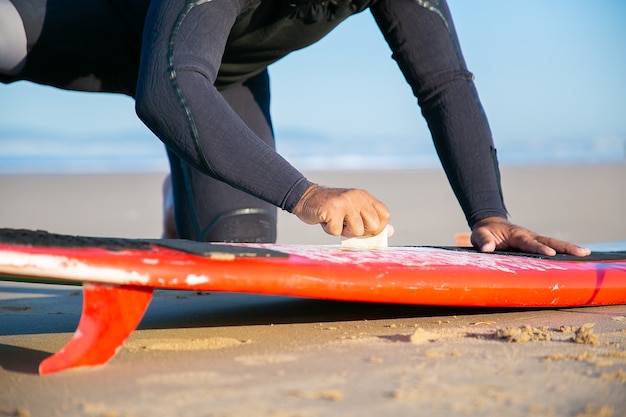 Бесплатное фото Мужчина-серфер в гидрокостюме, смазывая доску для серфинга на песке на берегу океана